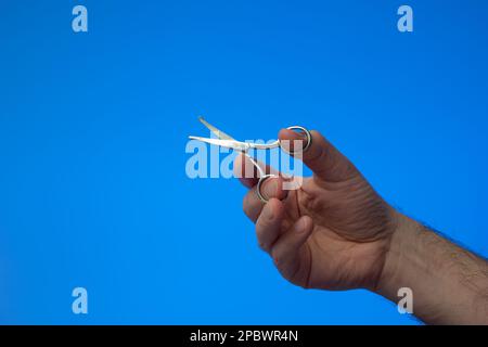 Small all metal scissor held in use by Caucasian male hand. Close up studio shot, isolated on blue background. Stock Photo