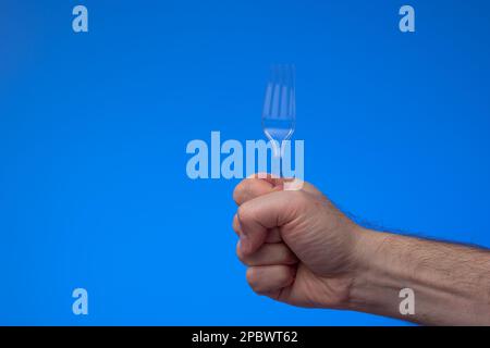 Transparent plastic single use fork held in hand by man. Close up studio shot, isolated on blue background. Stock Photo