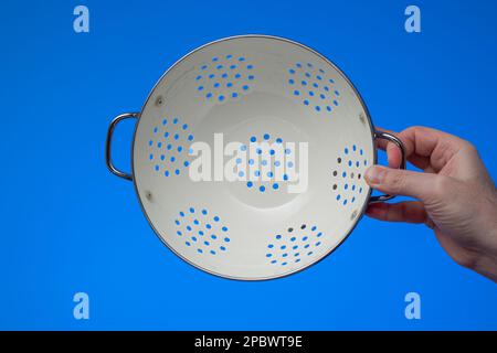 White metal round empty kitchen colander held by male hand. Close up studio shot, isolated on blue background. Stock Photo