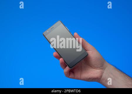 Generic black smartphone held by male hand. Close up studio shot, isolated on blue background. Stock Photo