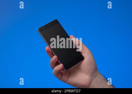 Generic black smartphone held by male hand. Close up studio shot, isolated on blue background. Stock Photo