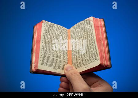 Old vintage small opened Bible book held in hand by Caucasian male. Close up studio shot, isolated on blue background. Stock Photo