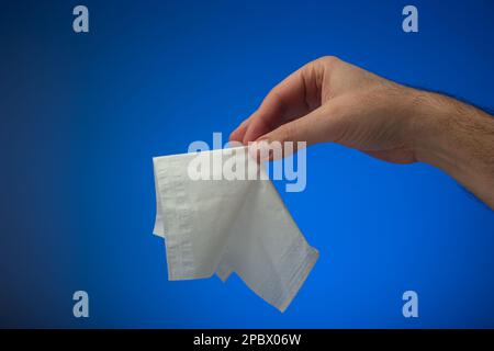 White paper napkin or tissue held in hand by Caucasian male. Close up studio shot, isolated on blue background. Stock Photo
