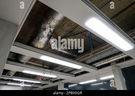 Large pipes mounted on the ceiling of an industrial building corridor. Removed panels to ease access. Neon lights, interior shot, no people.. Stock Photo
