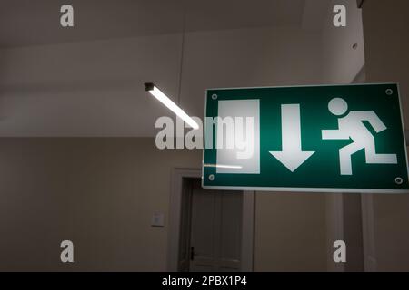 Green illuminated emergency exit sign, wall mounted inside building corridor. Defocused door in the background, no people. Stock Photo