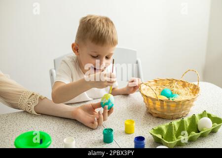 Happy child with mom coloring eggs. There are paints and a wicker basket on the table. Easter Stock Photo