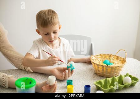 A child and his mother paint eggs. There are paints and a wicker basket on the table. Easter Stock Photo