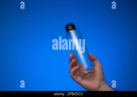 Empty transparent plastic water bottle with cap held in hand by Caucasian male hand. Close up studio shot, isolated on blue background. Stock Photo