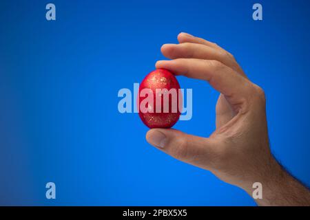 Hand painted and decorated red easter egg held in hand by Caucasian male. Close up studio shot, isolated on blue background Stock Photo