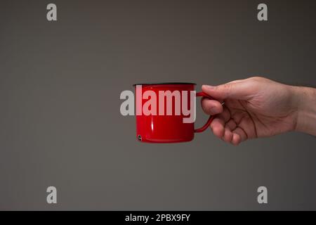 Red enameled tin camping cup or mug held in hand by a Caucasian male. Close up studio shot, isolated on gray background. Stock Photo