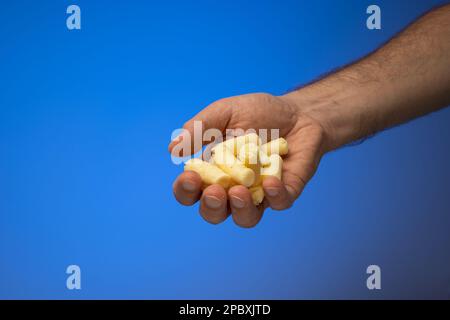 Handfull of  golden corn puffs held in hand by Caucasian male hand. Close up studio shot, isolated on blue background. Stock Photo