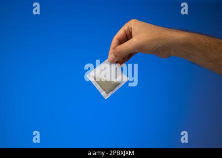 Single white paper tea bag held in hand by Caucasian male hand. Close up studio shot, isolated on blue background. Stock Photo