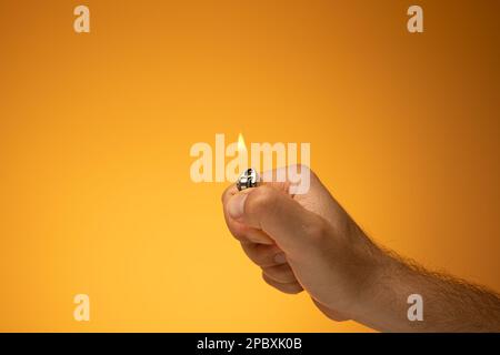 Lit cigarette lighter and flame held in hand by Caucasian male hand. Close up studio shot, isolated on orange background. Stock Photo