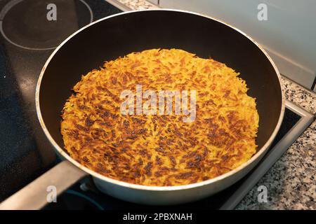 Homemade Swiss potato rosti or Rösti in a large frying pan on an electric stove. Top side view, no people. Stock Photo