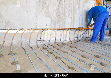A worker in a blue uniform is engaged in laying a floor heating cable on concrete, a man from the back Stock Photo