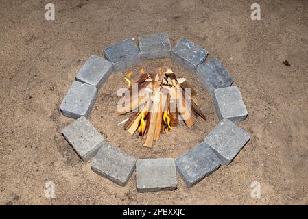 Prepared chopped pieces of wood to make a fire in a stone circle on the sand, visible flames. Stock Photo