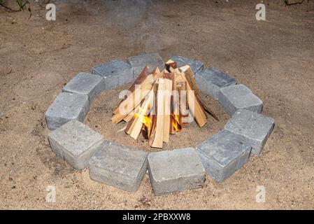 Prepared chopped pieces of wood to make a fire in a stone circle on the sand, visible flames. Stock Photo