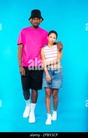 Full length of stylish young multiracial couple in casual outfits and white sneakers cuddling and looking at camera against blue background Stock Photo