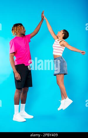 Side view of positive young ethnic female in casual clothes jumping for giving high five to tall African American boyfriend against blue background Stock Photo