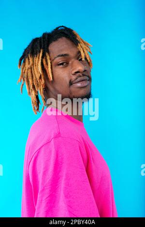 Side view of self assured young African American male model with dreadlocks in trendy outfit standing against blue background looking at camera Stock Photo