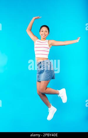 Full length of excited young mixed race female model with dark short hair in top and denim skirt smiling and looking at camera while jumping against b Stock Photo
