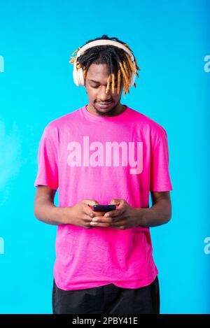 Young African American guy in pink t shirt listening to favorite song on headphones while browsing mobile phone against blue background Stock Photo