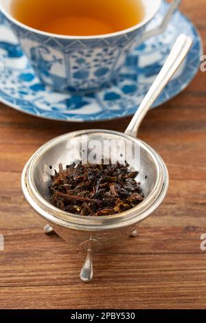 Antique old fashion silver tea strainer and stand with wet tea leaves in front of a cup of tea on a wooden table Stock Photo