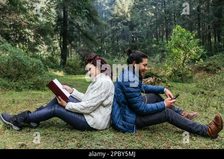 Full body side view of positive adult woman in warm knitted sweater reading book while sitting on ground near bearded man in jacket listening to music Stock Photo