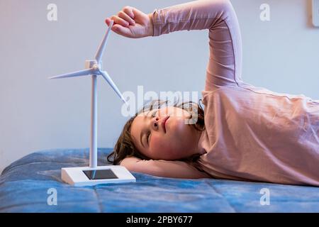 Positive cute preteen girl with curly brown hair in pink sweater lying on soft bed and spinning vane on plastic model of wind generator in bedroom Stock Photo