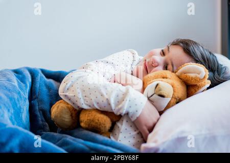 Cute preteen girl in pajamas sleeping on soft white pillow and embracing soft toy Stock Photo