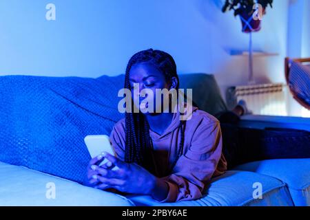 Focused young African American female in casual clothes browsing mobile phone while lying on sofa in blue neon light Stock Photo