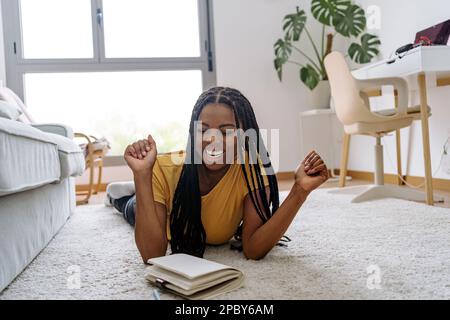 Positive young African American female in casual t shirt with long braids smiling and writing ideas in copybook while lying on carpet against window Stock Photo