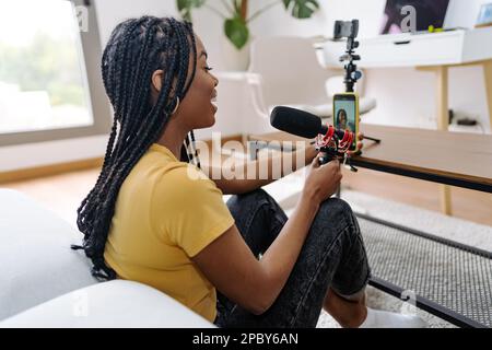 Side view of positive young African American female blogger in casual clothes with braids smiling while recording voice broadcast with microphone and Stock Photo