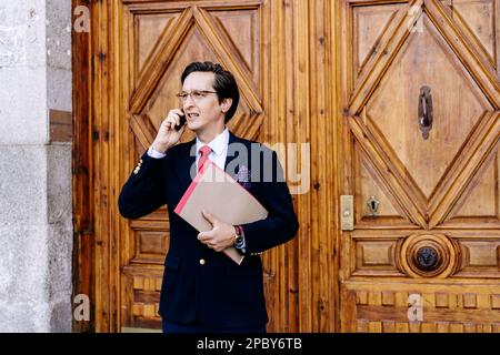 Busy entrepreneur in glasses standing with documents against building door and speaking on mobile phone while looking away Stock Photo