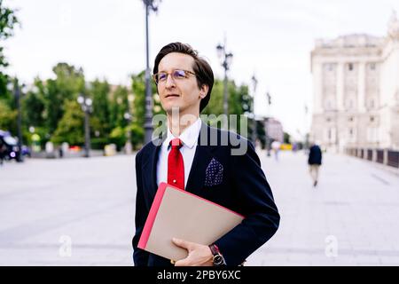Respectable man in formal clothes standing with documents and looking away on blurred background of city street Stock Photo
