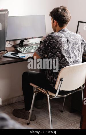 Back view full body of unrecognizable male freelancer in black t shirt with white flowers sitting on co comfortable chair at wooden desk and using com Stock Photo