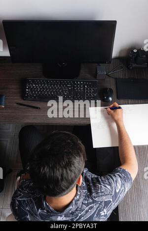 Top view of anonymous male freelancer in black t shirt sitting at wooden table near computer and writing on white paper Stock Photo