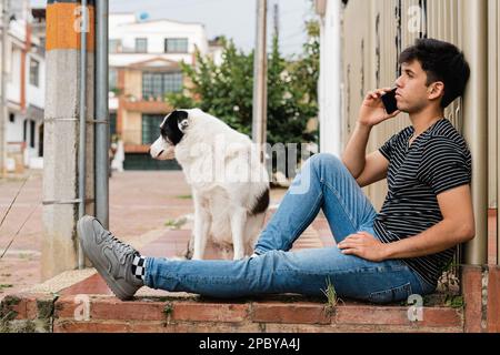 Full body side view of concentrated young male owner in casual clothes sitting on brick stairs and talking via smartphone while looking away pensively Stock Photo