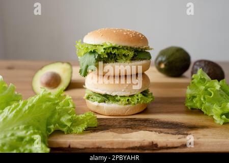 Tasty vegetarian burgers with fresh lettuce and avocado on wooden cutting board against gray background Stock Photo