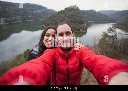 Portrait of smiling couple in warm clothes looking at camera and taking selfie against lake surrounded with mountains during hiking Stock Photo