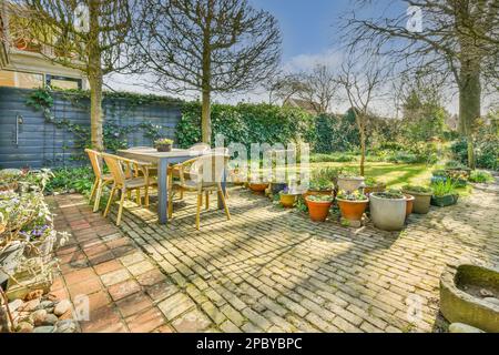 Wooden table with chairs and green potted plants placed on terrace of cafe in sunny day at backyard beside green lush garden Stock Photo