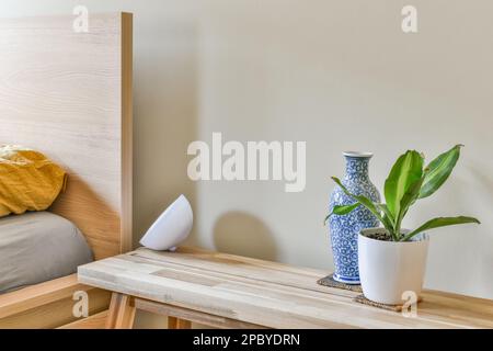 Interior design of nightstand near headboard of wooden bed in light bedroom decorated with green potted houseplant and blue vase Stock Photo