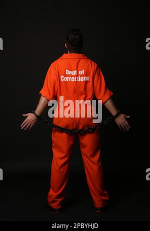 Prisoner in jumpsuit with chained hands on black background, back view Stock Photo