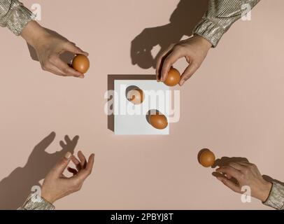 Four female hands reaching for fresh brown chicken eggs on white rectangle stand.  Minimal food concept. Flat lay. Stock Photo