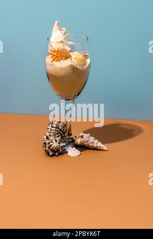 transparent glass filled with sand decorated with various sized conches and starfish placed on beige table near blue wall Stock Photo