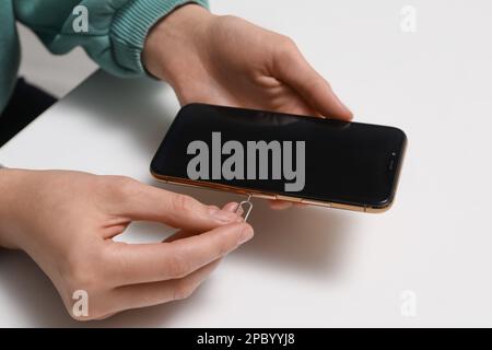 Woman with ejector opening SIM card tray in smartphone at white table, closeup Stock Photo