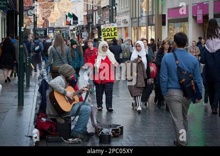 Shoppers of all kinds stroll Dublin's Grafton Street, and two Muslim women walk past a guitarist musician playing for tips. Stock Photo
