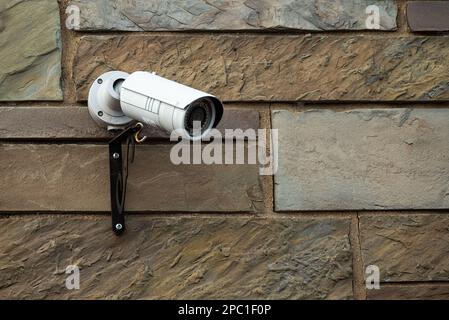 A white video surveillance camera mounted high on a rock faced exterior wall of a building in Upstate NY, USA Stock Photo
