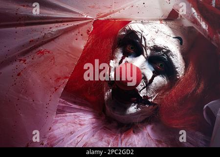 Terrifying clown staring through hole in torn bloodstained plastic film, closeup. Halloween party costume Stock Photo