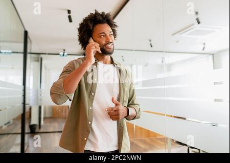 Male office employee having phone conversation, connecting with customers. Confident African-American man in smart casual wear talking on the smartphone while walkiing at the coworking office Stock Photo